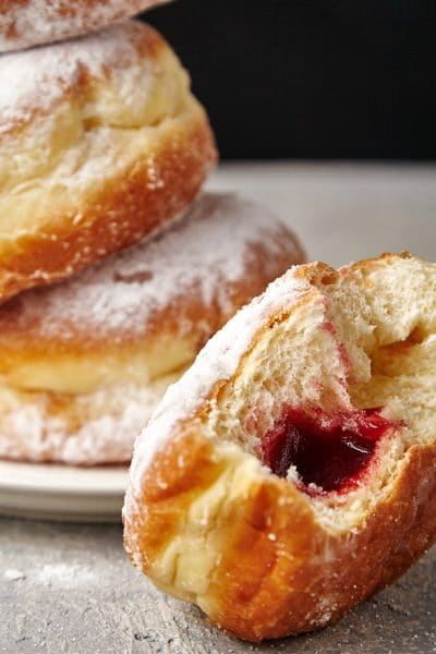 A stack of jelly doughnuts on a plate in front of a half eaten jam filled doughnut